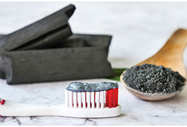 Charcoal toothpaste on a toothbrush next to bricks of charcoal and ground charcoal on a wooden spoon